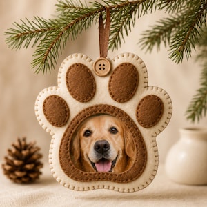 Puede incluir: Un adorno de fieltro con forma de pata que presenta una base color crema con almohadillas marrones y un recorte central que muestra la foto de un golden retriever. El adorno cuelga de una cinta marrón sujeta a un pequeño botón de madera.