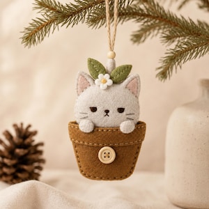 May include: A felt hanging ornament featuring a light grey cat with pink-lined ears peeking out of a brown felt flower pot. The cat has a small white flower and two green leaves on its head. A light-coloured wooden button is centred on the pot.