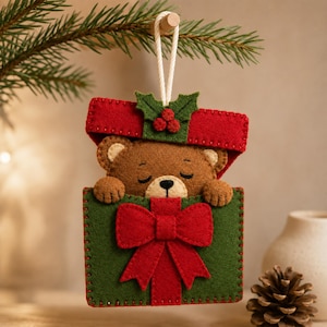 May include: A felt Christmas ornament featuring a brown bear peeking out of a green gift box with a red bow. The lid of the box is red with green holly leaves and three red berries. The ornament hangs from a white cord and has visible stitching.