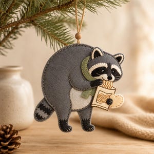 Peut inclure: Une décoration en feutrine représentant un raton laveur gris, blanc et noir, portant une écharpe vert sauge. Le raton laveur tient un sac en papier beige contenant des nouilles et un petit biscuit en forme de cœur, suspendu par une ficelle.