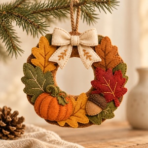 Peut inclure: Une décoration circulaire en feutrine ornée de motifs automnaux, incluant des feuilles orange, jaunes, vertes et rouges, une petite citrouille orange et un gland marron. Un nœud en feutrine crème brodé de feuilles surmonte le tout avec une boucle.