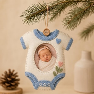 May include: A felt ornament shaped like a baby onesie with blue trim, a wooden button at the top, and a central oval photo frame containing a picture of a swaddled infant. The ornament features a small blue heart and a pink flower appliqué.