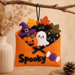 Op de afbeelding: Een oranje vilten envelopornament met het woord Spooky in zwarte letters. De envelop bevat een paarse heksenhoed, een oranje pompoen, een wit spookje, een zwarte vleermuis, een zwarte kat en herfstbladeren. Een zwart strikje is bevestigd.