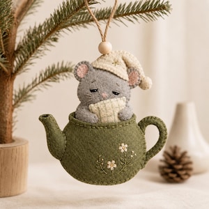 Op de afbeelding: Een vilten hangornament met een grijze muis die een crèmekleurige slaapmuts draagt en een klein kussentje vasthoudt, genesteld in een donker olijfgroene theepot versierd met geborduurde witte bloemen, opgehangen aan een bruin touwtje.