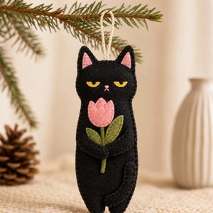 Puede incluir: Un adorno de fieltro negro con forma de gato, ojos amarillos, orejas rosas y nariz rosa. El gato sostiene un tulipán rosa con tallo y hojas verdes. El adorno presenta costuras negras visibles en los bordes y un lazo de cuerda blanca.