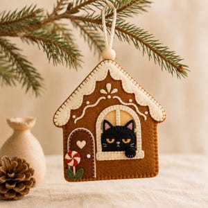 Può includere: Una decorazione in feltro marrone a forma di casetta di pan di zenzero con un gatto nero che guarda fuori dalla finestra. La casa presenta dettagli in glassa bianca, un piccolo cuore sulla porta e una decorazione a caramella menta.
