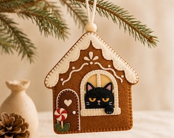 May include: A brown felt gingerbread house ornament featuring a black cat looking out of a window. The house has white icing details, a small heart on the door, and a red and white peppermint candy decoration. It hangs from a white cord.