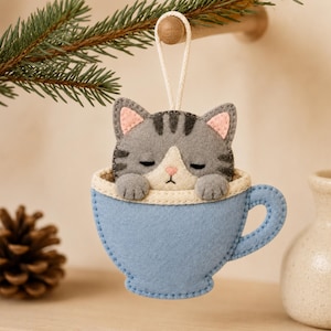 Peut inclure: Une décoration en feutrine représentant un chat tigré gris avec des rayures foncées et un museau blanc, installé dans une tasse à thé bleu clair. Le chat a les yeux fermés, l'intérieur des oreilles rose et un petit nez rose. Une boucle en corde blanche est fixée au sommet.