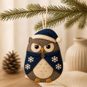 May include: A felt owl ornament featuring a dark blue Santa hat with a cream trim, a dark blue cape with white embroidered snowflakes, a small brown button, and a cream-colored belly. The owl is gray with orange feet and a yellow beak.