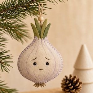 May include: A felt onion ornament featuring a pale lavender and white body with a sad facial expression, green felt leaves at the top, and a small wooden bead on a jute hanging loop. The onion has visible stitching along the edges.