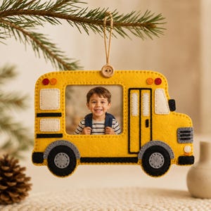 May include: A yellow felt school bus ornament featuring a central cutout window displaying a photograph of a smiling child. The bus has black wheels, white window accents, and a small wooden button at the top where a jute twine loop is attached.