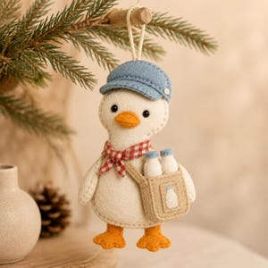 May include: A felt hanging ornament shaped like a white duck wearing a blue cap and a red and white checkered scarf. The duck carries a tan satchel containing two small white bottles, with a milk bottle icon stitched onto the front of the bag.