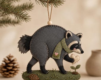 May include: A felt raccoon ornament in shades of charcoal and light gray, wearing a sage green scarf with a small pink heart detail. The raccoon holds a cream-colored heart while standing on a green felt patch with a sprout and a brown leaf.