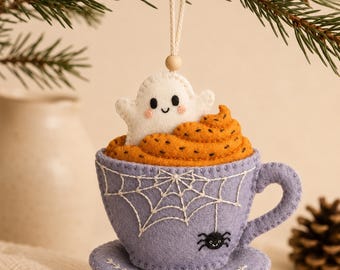 May include: A felt hanging ornament shaped like a lavender teacup and saucer. The cup features white embroidered spiderwebs and a small black spider. Inside the cup is orange whipped topping with a small white ghost with pink cheeks on top.
