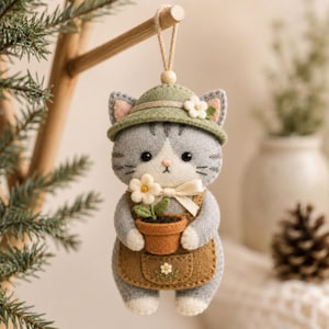 May include: A felt cat ornament featuring a grey and white tabby cat wearing a green hat with a small white flower. The cat wears a brown apron with a small embroidered flower and holds a tiny brown potted plant with a white flower.