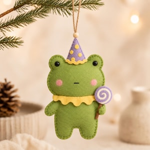 May include: A green felt frog ornament wearing a purple party hat with yellow polka dots and a yellow ruffled collar. The frog has pink cheeks, black eyes, and holds a small purple and white swirl lollipop. It hangs from a brown twine string.