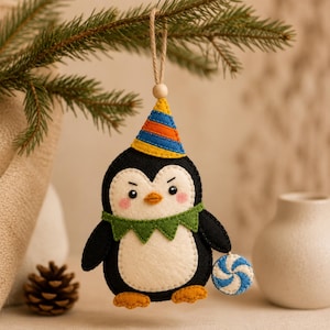 Puede incluir: Un adorno de fieltro con forma de pingüino, cuerpo blanco y negro, pies naranjas y un gorro de fiesta a rayas amarillas, azules y naranjas. El pingüino lleva un cuello verde festoneado y sostiene un caramelo de menta azul y blanco.