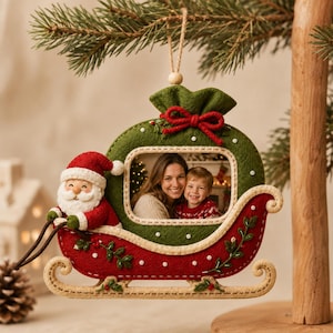 May include: A felt Christmas ornament shaped like a red and green sleigh with a rectangular photo frame in the centre. A small Santa Claus figure sits at the front of the sleigh, which is decorated with white stitching, green leaf embroidery, and a red bow.