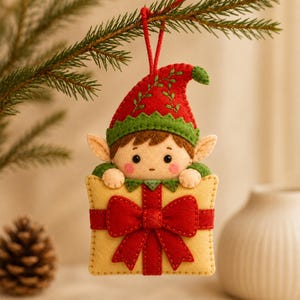 Puede incluir: Un adorno navideño de fieltro con forma de elfo asomándose sobre un regalo amarillo. El elfo lleva un gorro rojo con borde verde y bordados. El regalo está envuelto con un gran lazo de fieltro rojo. Tiene una cuerda roja en la parte superior.