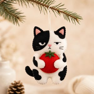 May include: A felt cat ornament featuring a black and white patterned coat, pink ears, and a grumpy facial expression. The cat is holding a bright red tomato with a green stem in its paws. A thin white string is attached to the top for hanging.