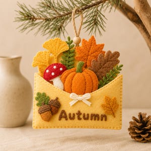 May include: A felt envelope ornament featuring the word Autumn in brown stitching. The envelope is filled with a felt orange pumpkin, a red and white mushroom, yellow ginkgo leaves, orange maple leaves, brown oak leaves, and green ferns.
