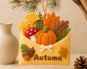 May include: A felt envelope ornament featuring the word Autumn in brown stitching. The envelope is filled with a felt orange pumpkin, a red and white mushroom, yellow ginkgo leaves, orange maple leaves, brown oak leaves, and green ferns.