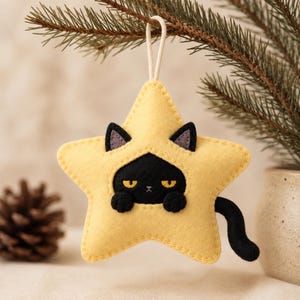 Puede incluir: Un adorno de fieltro en forma de estrella de color amarillo pálido con un gato negro asomándose desde el centro. El gato tiene ojos amarillos, orejas pequeñas y dos patas apoyadas en el borde de la estrella. Una cola negra sobresale por el lado y tiene un cordón blanco.
