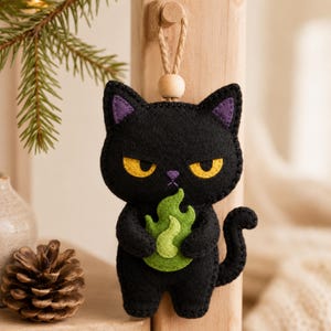 Puede incluir: Un adorno de fieltro negro con forma de gato, ojos amarillos, interior de orejas moradas y nariz morada. El gato sostiene una llama de color verde claro entre sus patas. Cuelga de un cordel de yute con una cuenta de madera en un poste.