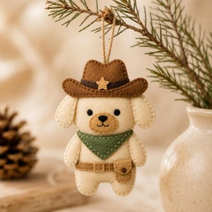 May include: A felt dog ornament wearing a brown cowboy hat with a small star, a green bandana, and a brown belt with a pouch. The cream-colored dog has black eyes and a stitched nose and mouth. It hangs from a thin twine loop for display purposes.