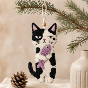 Peut inclure: Une décoration en feutrine en forme de chat blanc à taches noires, avec des oreilles roses et une expression boudeuse. Le chat tient un petit poisson violet clair entre ses pattes avant. Il est suspendu par un cordon blanc avec une perle en bois.