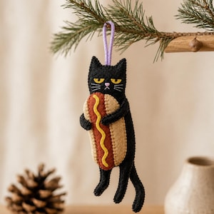 May include: A felt hanging ornament shaped like a black cat with yellow eyes and white whiskers, holding a tan hot dog with a brown sausage and a yellow mustard line. The cat has a small pink nose and is suspended by a light purple cord loop.