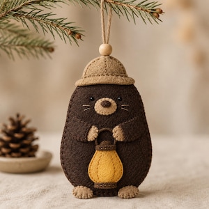 May include: A dark brown felt mole ornament wearing a tan hat and holding a yellow lantern. The figure has a light tan snout, small black eyes, and stitched details. It hangs from a thin twine loop with a small wooden bead at the top.