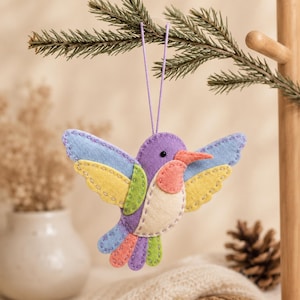 Puede incluir: Un adorno de fieltro con forma de colibrí que presenta cabeza y cuerpo morados, alas azul claro y amarillas, garganta color coral y cola multicolor. El ave está construida con costuras visibles en los bordes e incluye un cordón morado para colgar.
