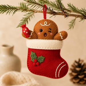Könnte beinhalten: Ein Weihnachtsanhänger aus Filz mit einem braunen Lebkuchenmännchen, das zwinkert und ein rotes Bonbon hält. Es steckt in einem roten Weihnachtsstrumpf mit weißem Umschlag, der mit grünen Stechpalmenblättern und roten Beeren verziert ist.