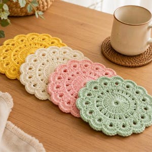 May include: A set of four round crochet coasters in yellow, cream, pink, and light green, each featuring a scalloped edge and a central circular pattern. The coasters are arranged in a staggered line on a wooden surface next to a ceramic mug.