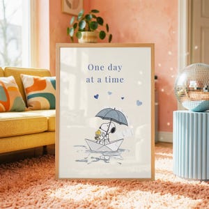 Pode incluir: Uma impressão artística emoldurada com fundo claro e o texto One day at a time em azul. A ilustração mostra Snoopy e Woodstock sentados num barco de papel branco sob um guarda-chuva azul escuro durante uma chuva leve com corações azuis.