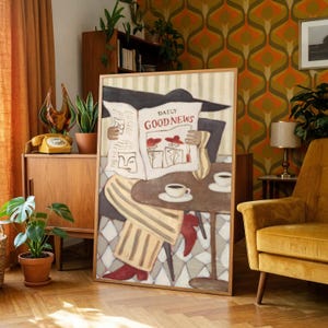 May include: A framed art print featuring a person sitting at a table reading a newspaper titled DAILY GOOD NEWS. The illustration shows a person wearing striped trousers and red boots, holding a newspaper with a coffee cup on the table in front.