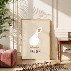 Pode incluir: Uma impressão de arte de parede retangular apresentando um ganso branco com bico e pés laranja, posicionado contra um fundo bege claro. O texto NICE BUM está impresso em letras maiúsculas pretas sem serifa abaixo da ilustração do ganso.