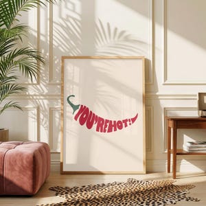 Pode incluir: Um poster retangular em moldura de madeira clara com o gráfico de uma pimenta vermelha. O texto "YOU'RE HOT!" está escrito numa fonte vermelha, negrito e sem serifa, seguindo a forma curva da pimenta sobre um fundo de cor creme.
