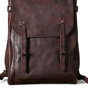 Könnte beinhalten: Ein dunkelbrauner Lederrucksack mit einer großen Umschlagklappe, die durch zwei vertikale Riemen mit Metallschnallen gesichert ist. Die Tasche hat eine glatte, matte Oberfläche mit sichtbaren Nähten an den Kanten und verstellbaren Seitenriemen.