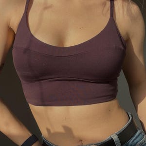 May include: A dark plum-colored cropped camisole top featuring thin shoulder straps, a scoop neckline, and a horizontal seam detail across the chest. The garment is paired with blue denim jeans secured by a black leather belt with a silver buckle.