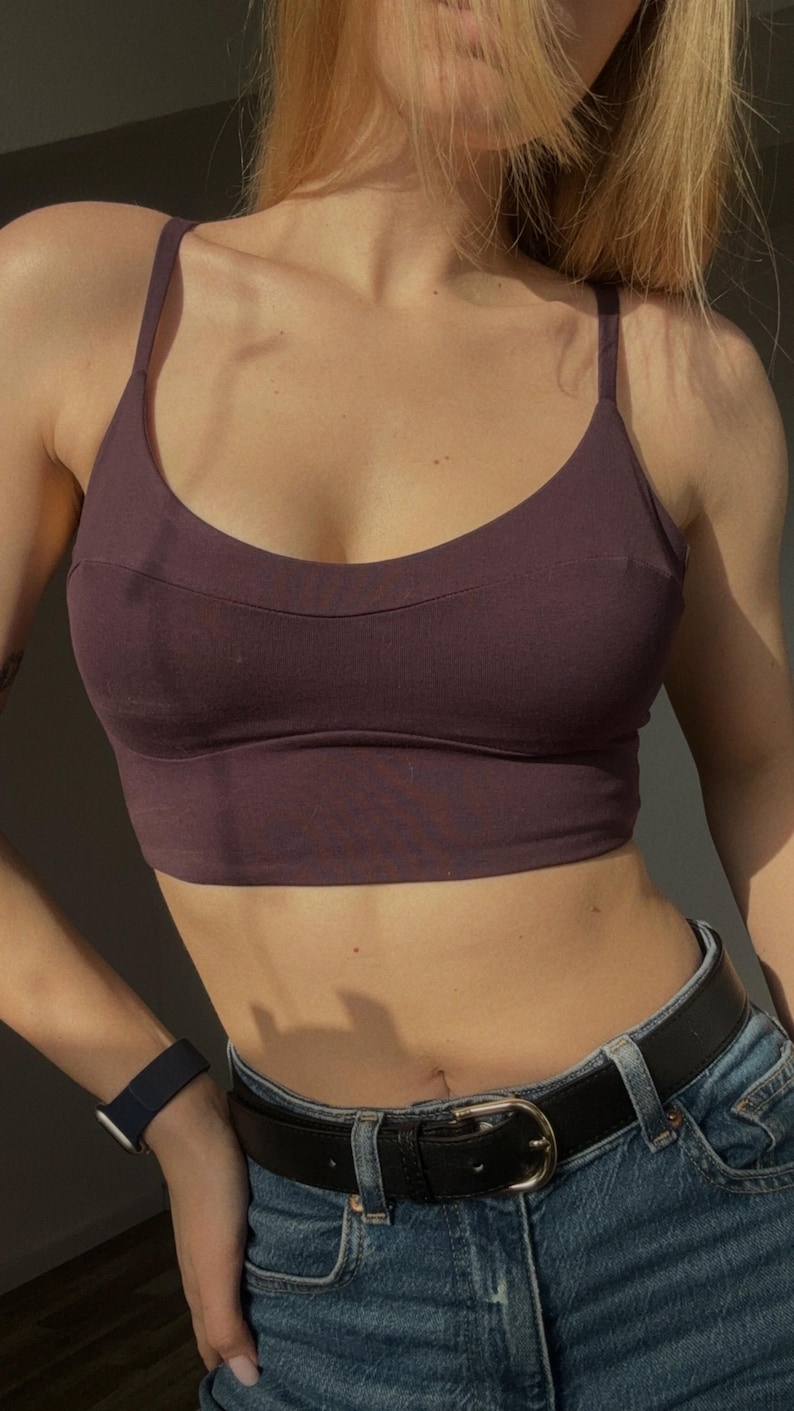 May include: A dark plum-colored cropped camisole top featuring thin shoulder straps, a scoop neckline, and a horizontal seam detail across the chest. The garment is paired with blue denim jeans secured by a black leather belt with a silver buckle.