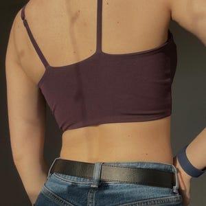 May include: A dark purple cropped camisole top with thin adjustable shoulder straps, paired with blue denim jeans featuring a black leather belt. The top has a straight back neckline and a fitted silhouette, worn with a blue wristband on the arm.