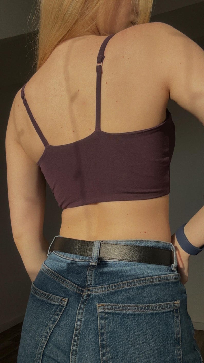 May include: A dark purple cropped camisole top with thin adjustable shoulder straps, paired with blue denim jeans featuring a black leather belt. The top has a straight back neckline and a fitted silhouette, worn with a blue wristband on the arm.