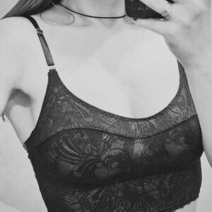 May include: A black lace bralette featuring a scoop neckline, thin adjustable shoulder straps with metallic hardware, and a wide band. The garment is crafted from a floral patterned lace fabric that extends from the neckline to the bottom hem.
