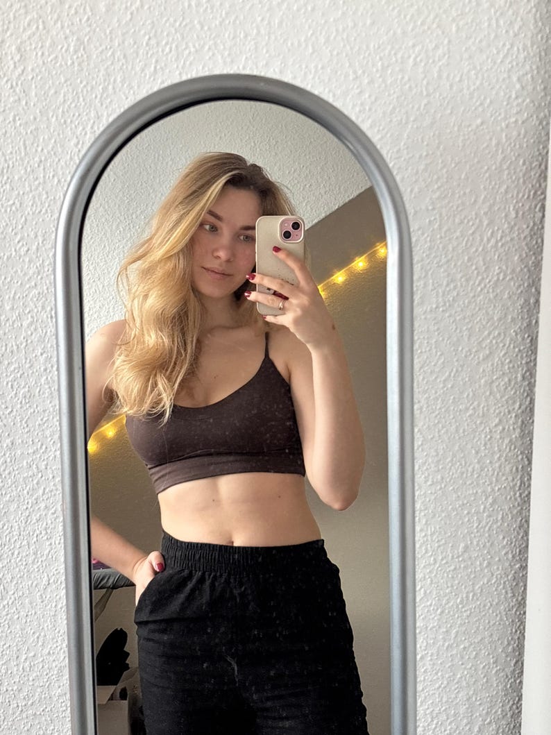 May include: A dark brown cropped camisole top with thin spaghetti straps and a scoop neckline, paired with black high-waisted pants featuring an elastic waistband. The garment is captured in a reflection within an arched silver-framed mirror.