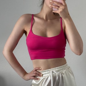 May include: A vibrant hot pink cropped tank top featuring thin spaghetti straps and a scoop neckline. The garment has a fitted silhouette with a horizontal seam detail across the chest area, providing a structured look for this sleeveless top.