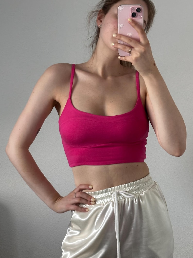 May include: A vibrant hot pink cropped tank top featuring thin spaghetti straps and a scoop neckline. The garment has a fitted silhouette with a horizontal seam detail across the chest area, providing a structured look for this sleeveless top.