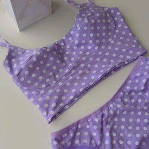 May include: A lavender two-piece set featuring a cropped top with thin straps and matching bottoms, both patterned with white polka dots. The set is labeled with the text Home cotton. A white rectangular box is positioned in the upper left corner.
