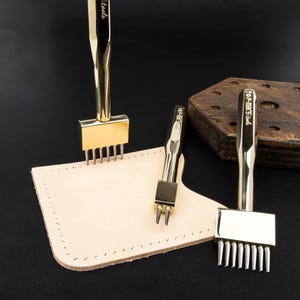 May include: A collection of gold-colored leatherworking tools, including stitching chisels and a two-prong tool, are displayed on a light tan leather piece. The tools are arranged on a dark background, suggesting a professional craft setting.